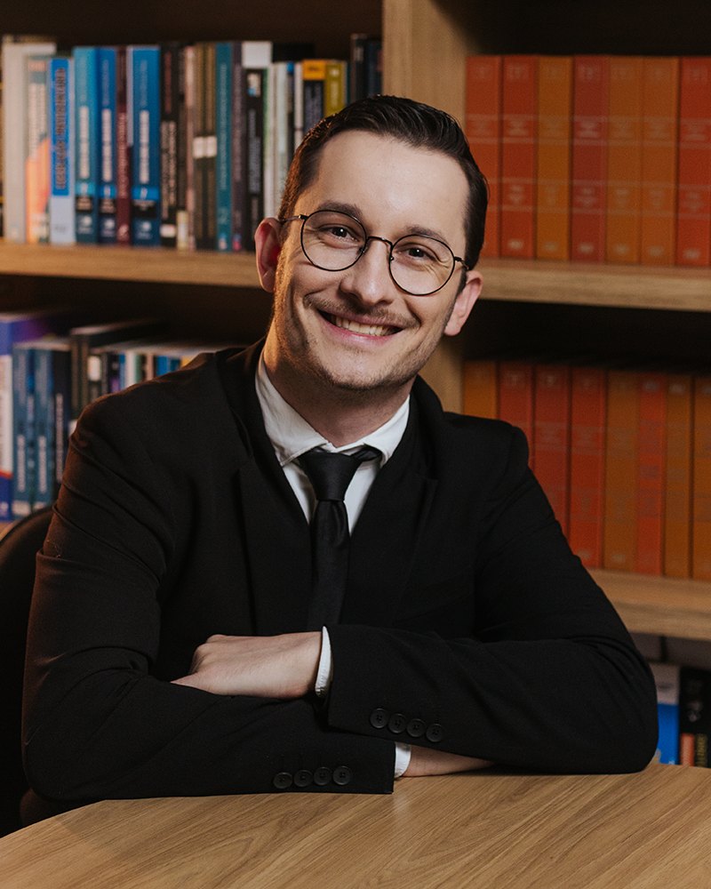 Thiago Goedert sentado à mesa, diante de uma estante de livros jurídicos